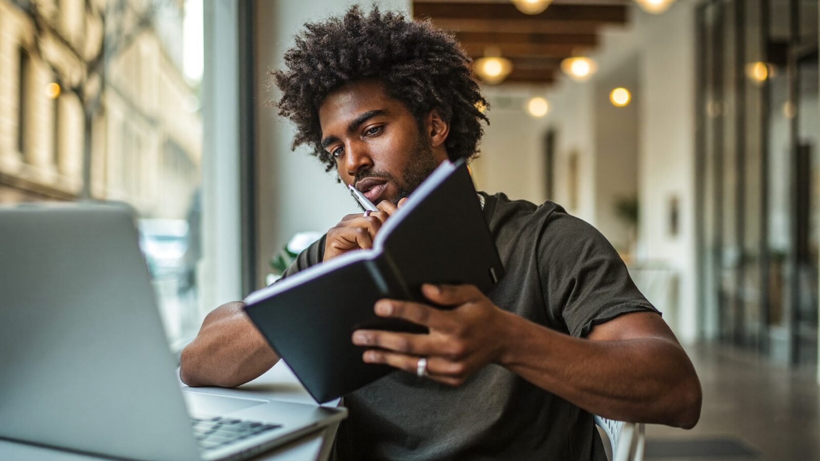 Man in his 30s with full curly hair thinking over a notebook at a cafe window
