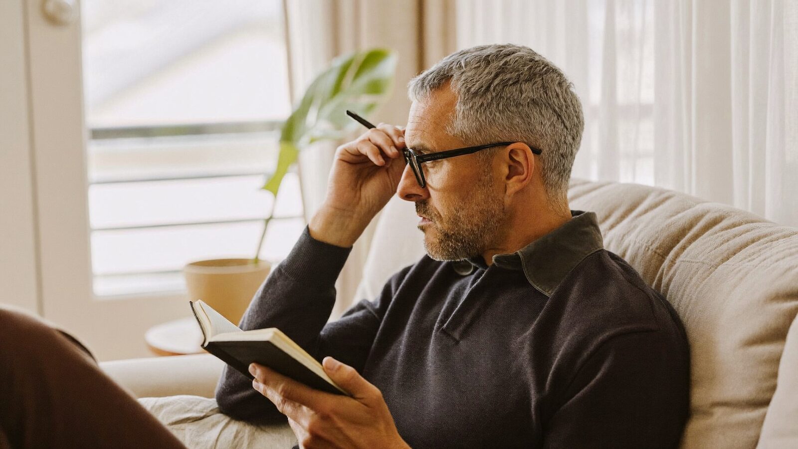 Man in his 40s with full grey hair sitting on a sofa with a notebook, thinking through his questions