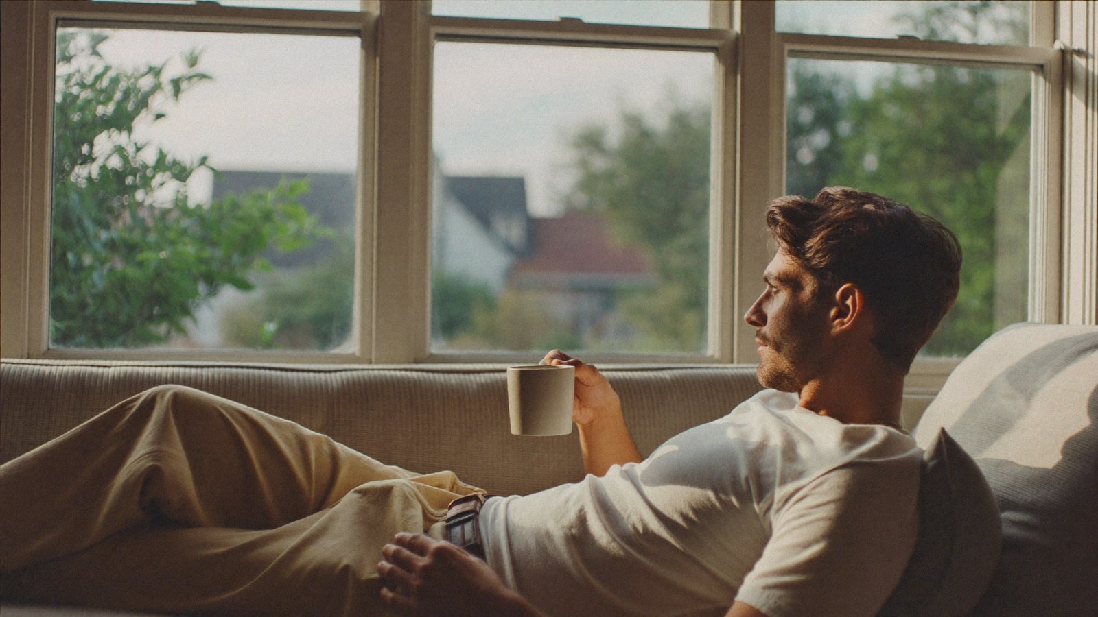 Man with thick dark hair resting on a sofa by a sunlit window with a mug of tea