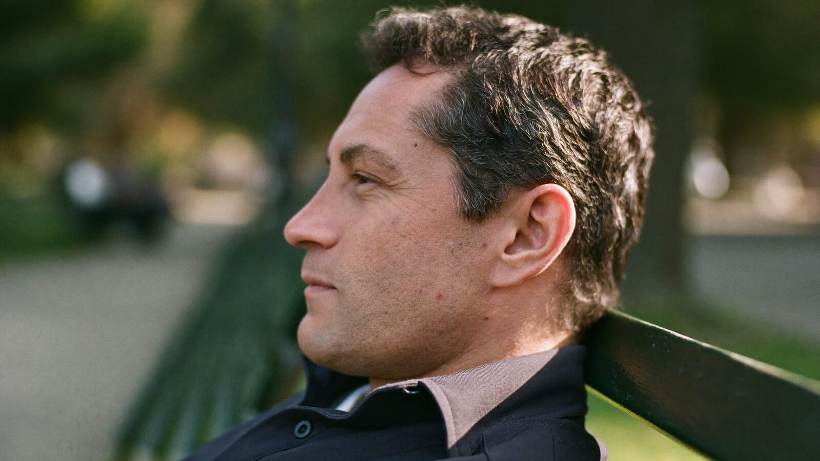 Relaxed man in his 40s with dark hair sitting on a park bench in afternoon light